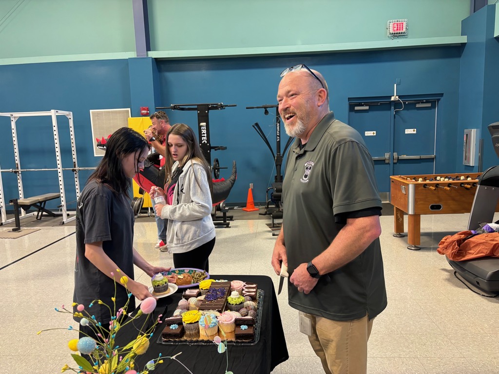 Three people in a gym, one man smiles at woman with flowers near a table with cupcakes.