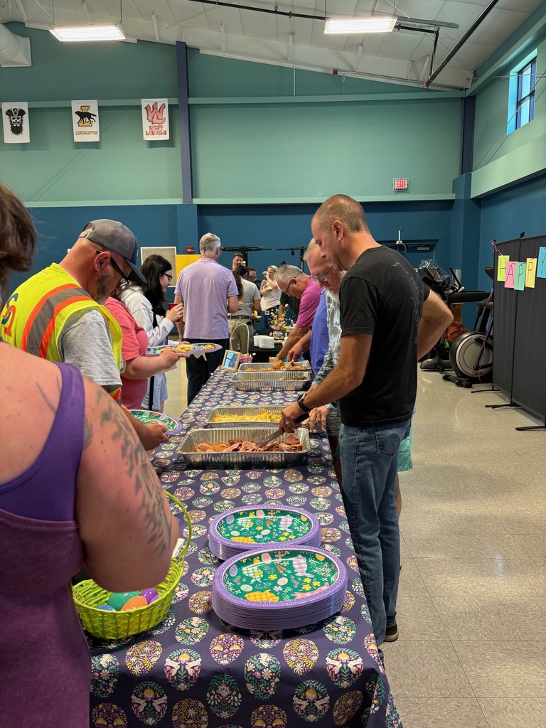 People gather around a long table with food at an indoor event, in a room with blue walls and exercise equipment.