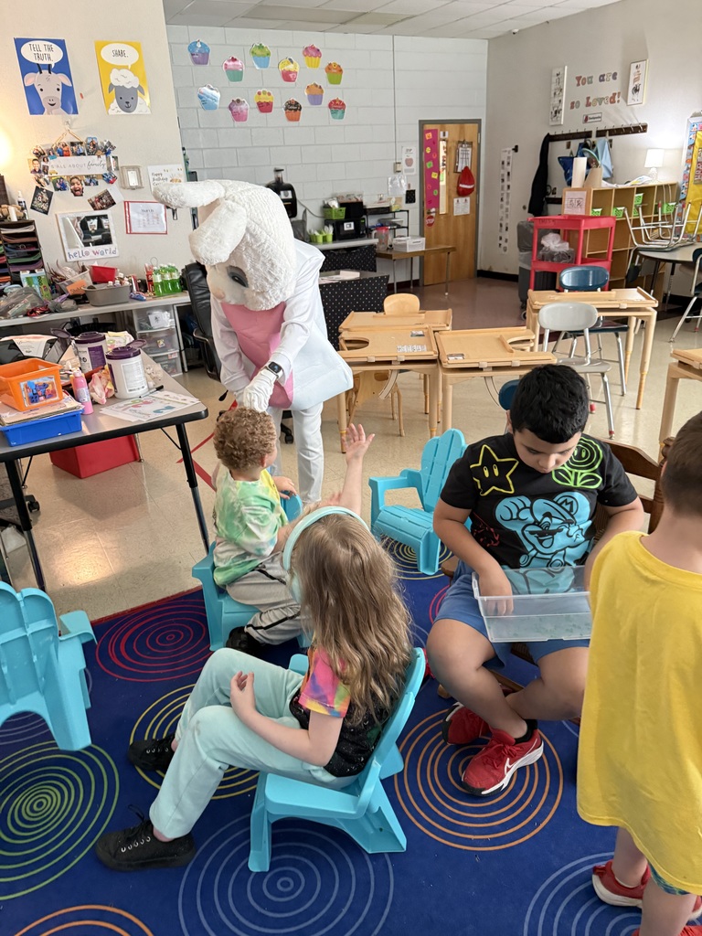 A classroom with kids sitting in chairs. A person in a bunny costume stands behind them.