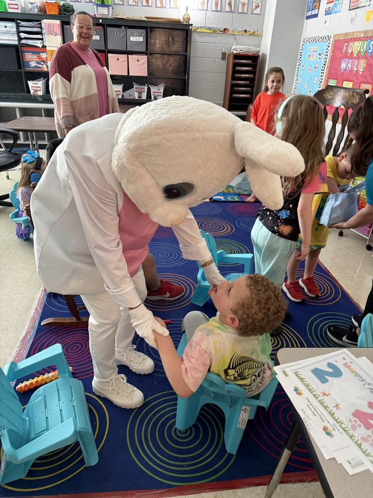 A mascot in a bunny costume shakes a child's hand in a classroom with chairs and tables.