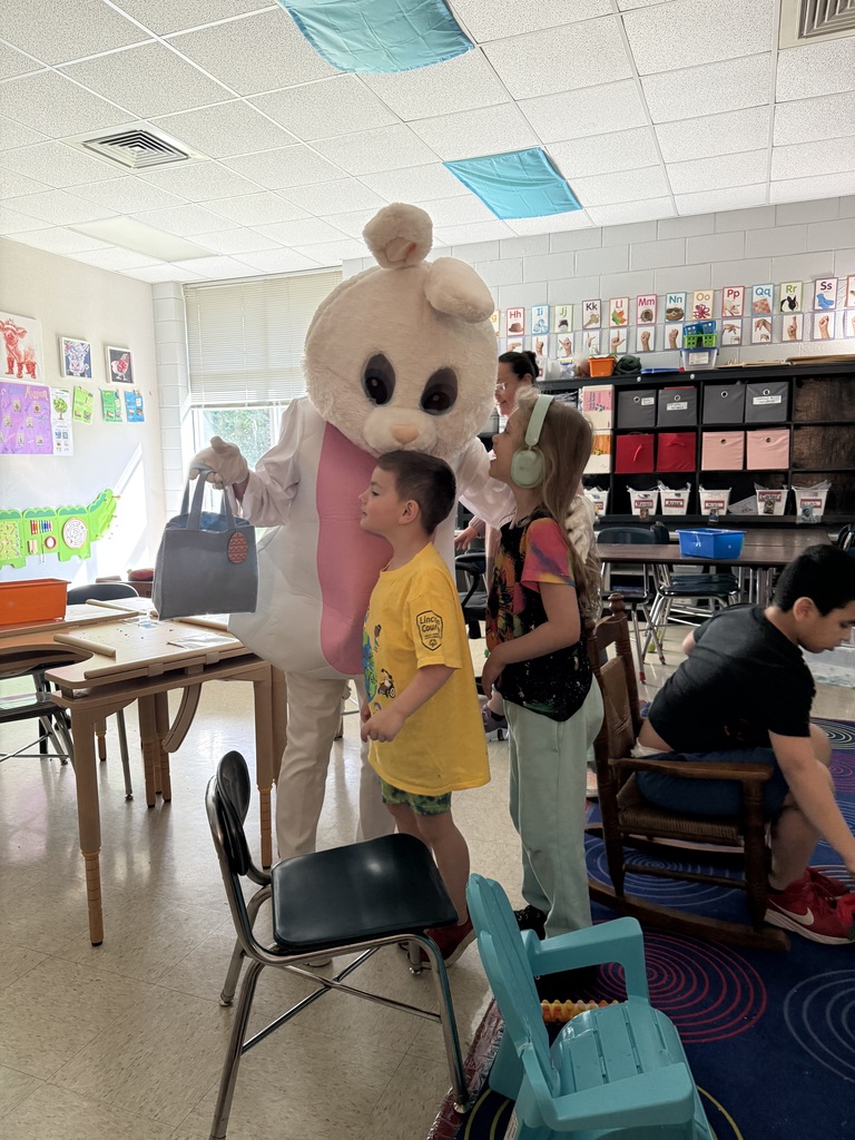 A bunny mascot wearing headphones interacts with a child and two others in a classroom.