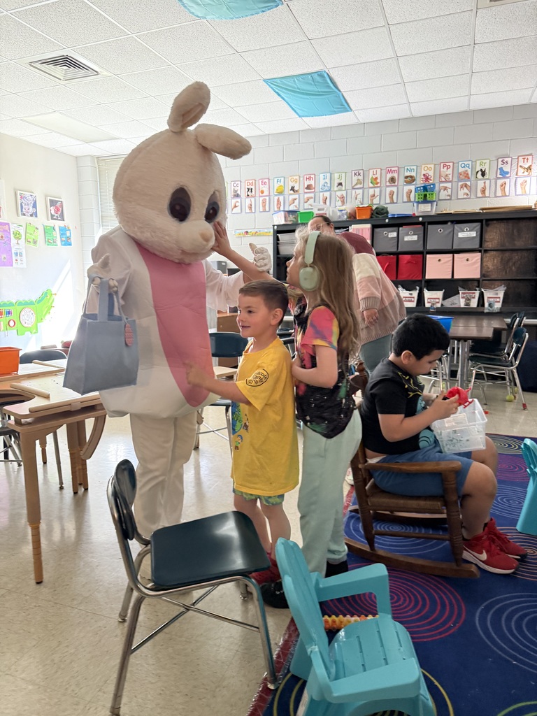 Children in a classroom interact with a mascot. A mascot wears a bunny costume. The children are standing and sitting on chairs.