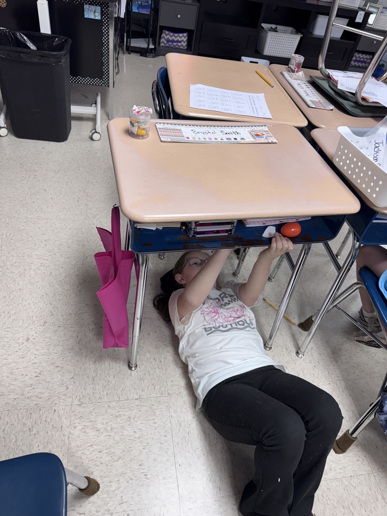 A student lies on the floor with their head under a desk in a classroom.
