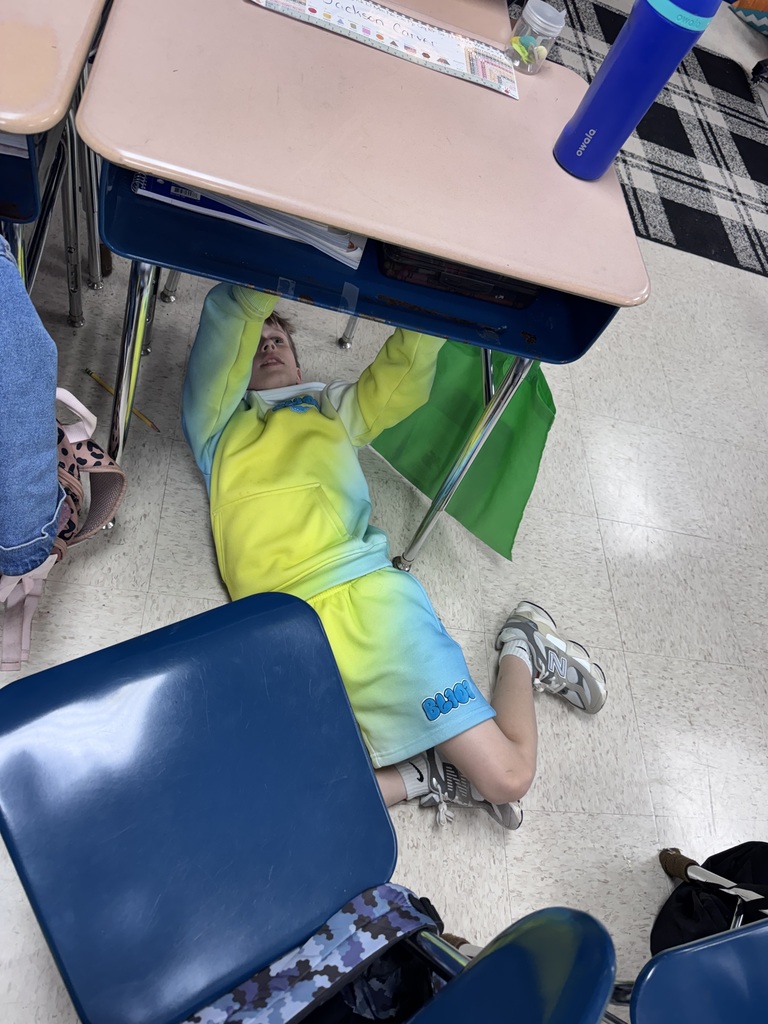 A person is lying on the floor under a desk, wearing a yellow and blue outfit. A chair is nearby.