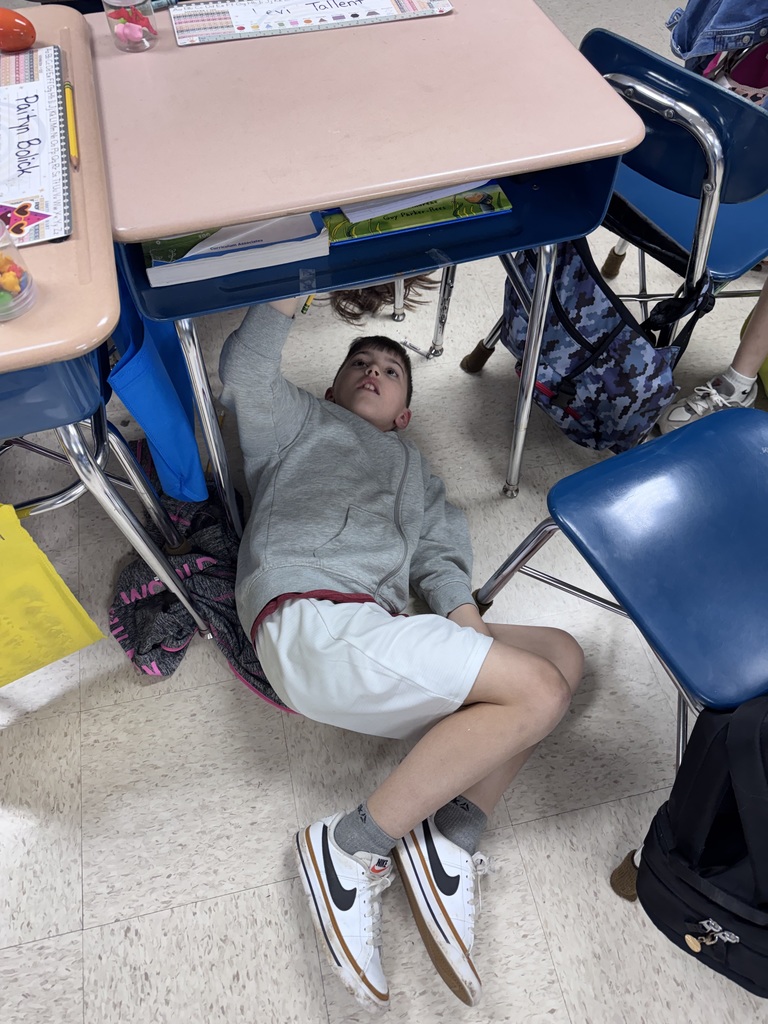 A person lies on the floor, head under a desk, in a classroom with blue chairs and desks.