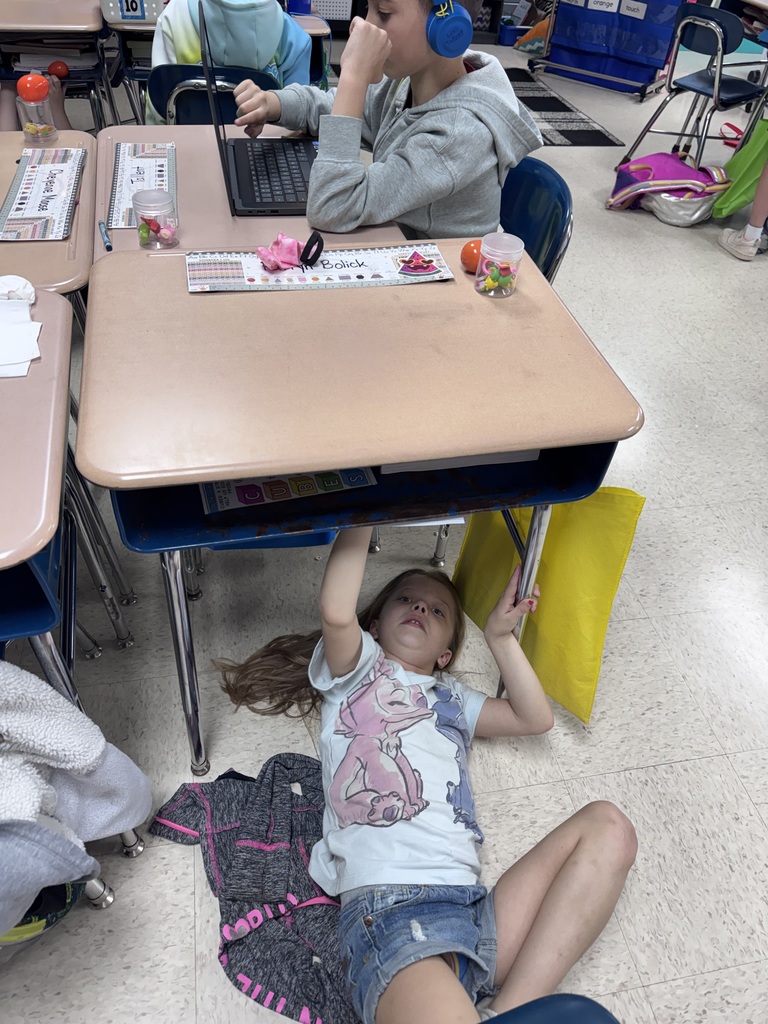 Two kids seated in chairs at a table, one of them lying on the floor under the desk.