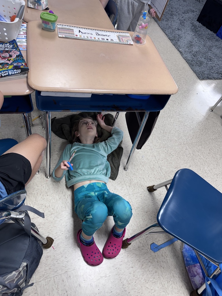 A child rests on the floor next to a desk, looking down. A backpack and two blue chairs are nearby.