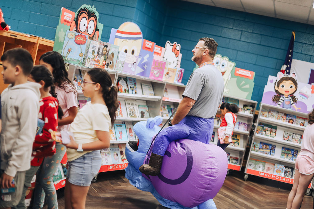 A man sits on a purple inflatable toy while surrounded by children in a colorful store.