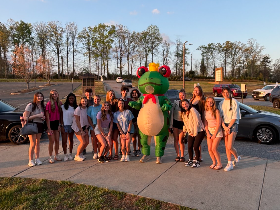 A group of young people and a frog mascot pose on a sidewalk in a parking lot.