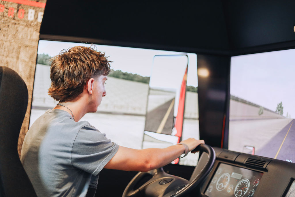 A person sits in a simulator, hands on the wheel, with three large screens showing a road ahead.