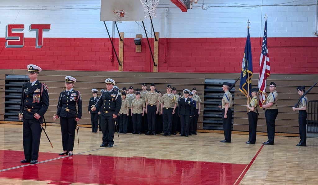 A group of uniformed people stand in a gymnasium, with flags behind them. One person holds a rifle.
