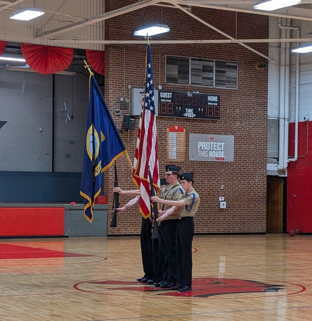 Four individuals standing in a gymnasium. One holds a flag. A basketball court is visible.