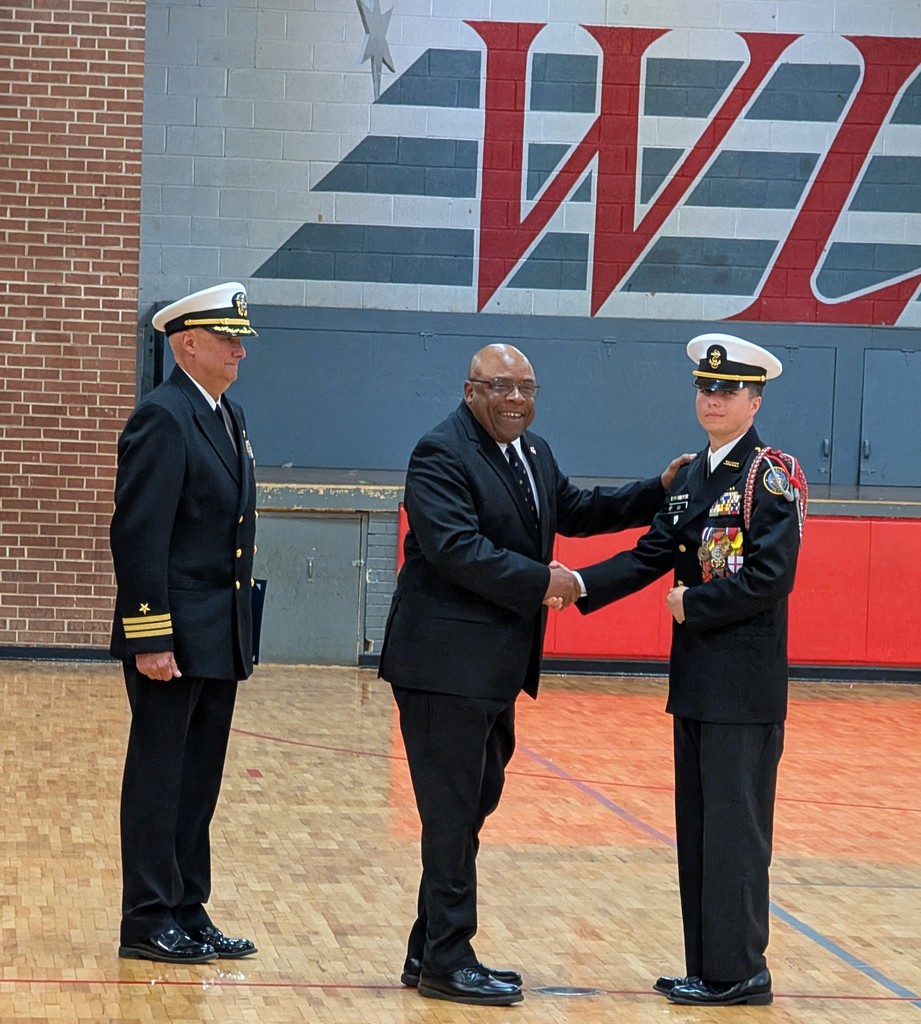 Three individuals in suits and hats stand on a basketball court, shaking hands. A brick wall with a large red "W" is behind them.