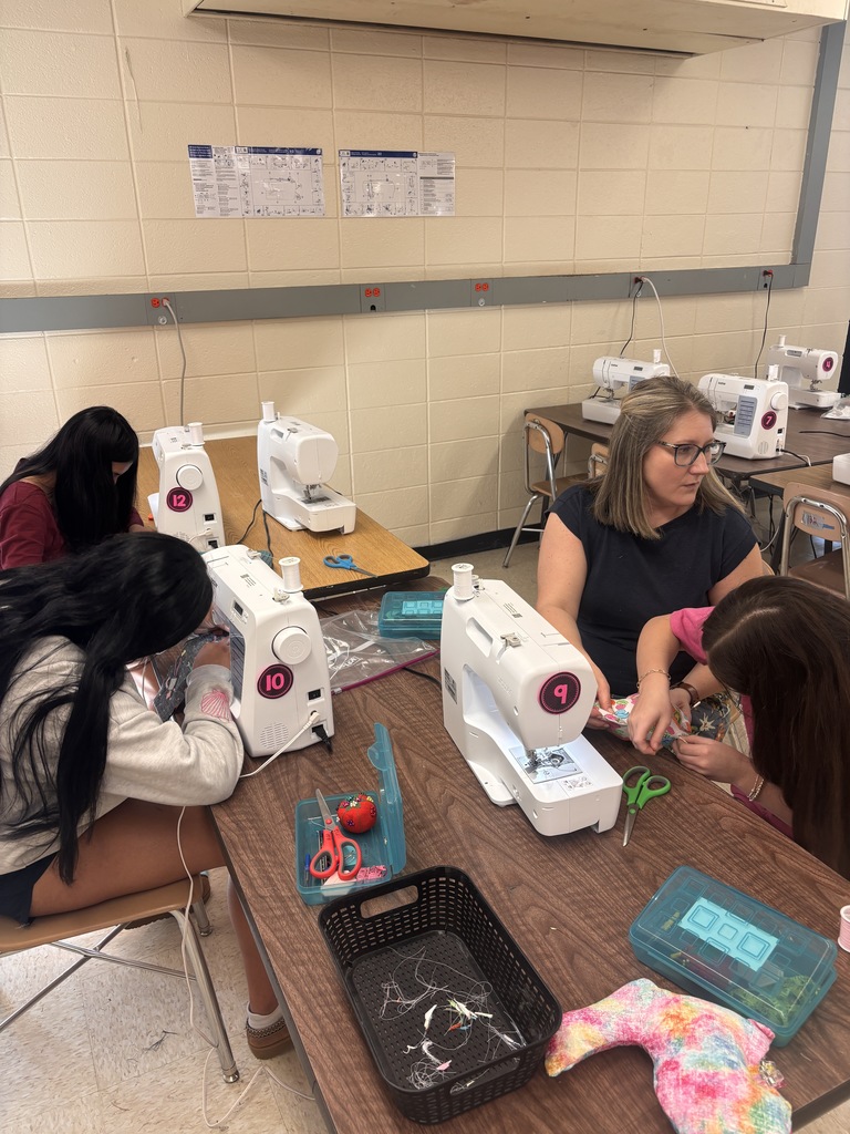 Four people are seated around a table in a classroom. Each person is using a sewing machine.