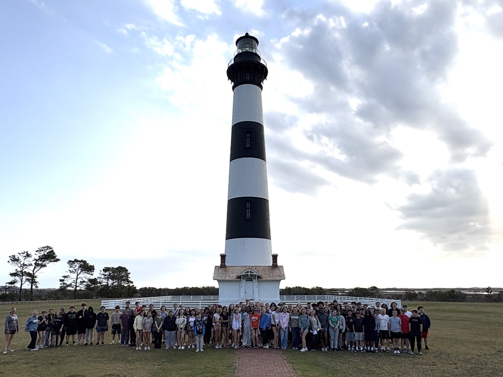 Group of people standing in front of the Cape Hatteras Lighthouse. A cloudy sky is above them.