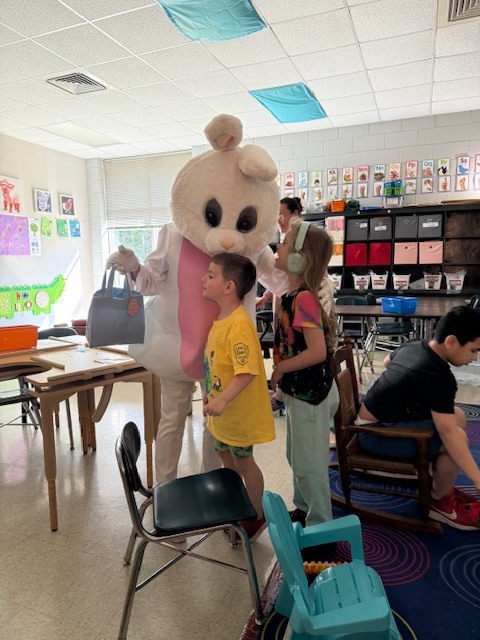 A mascot resembling a bunny stands in a classroom with children. Two children look at the mascot.