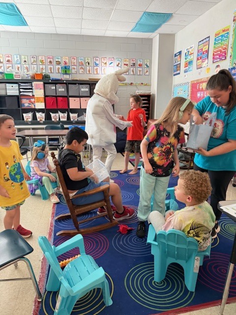 A classroom with a carpet, children, and an adult dressed in a white costume. A few kids sit in chairs while others stand.