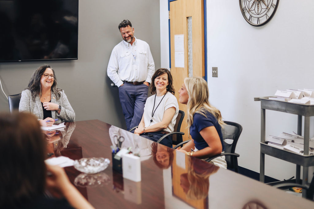 People seated around a conference table, one man standing, others engaged in conversation, with a TV and clock on the wall.