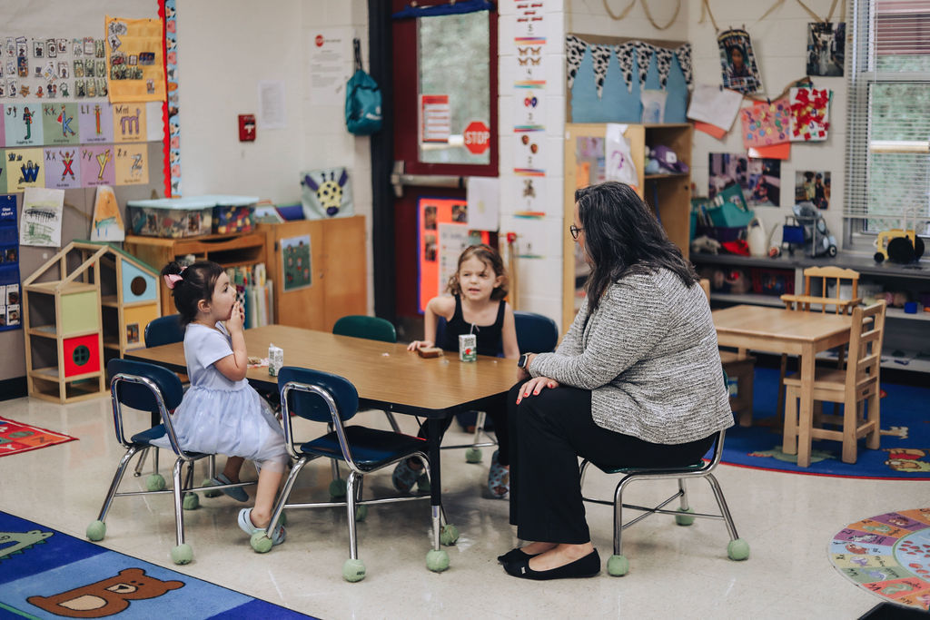 A woman and two young girls seated around a table in a classroom, with colorful decor and educational posters in the background.