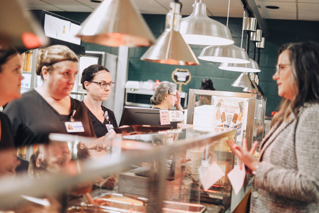 People inside a deli counter, with three women standing behind the counter, one with glasses and a name tag.