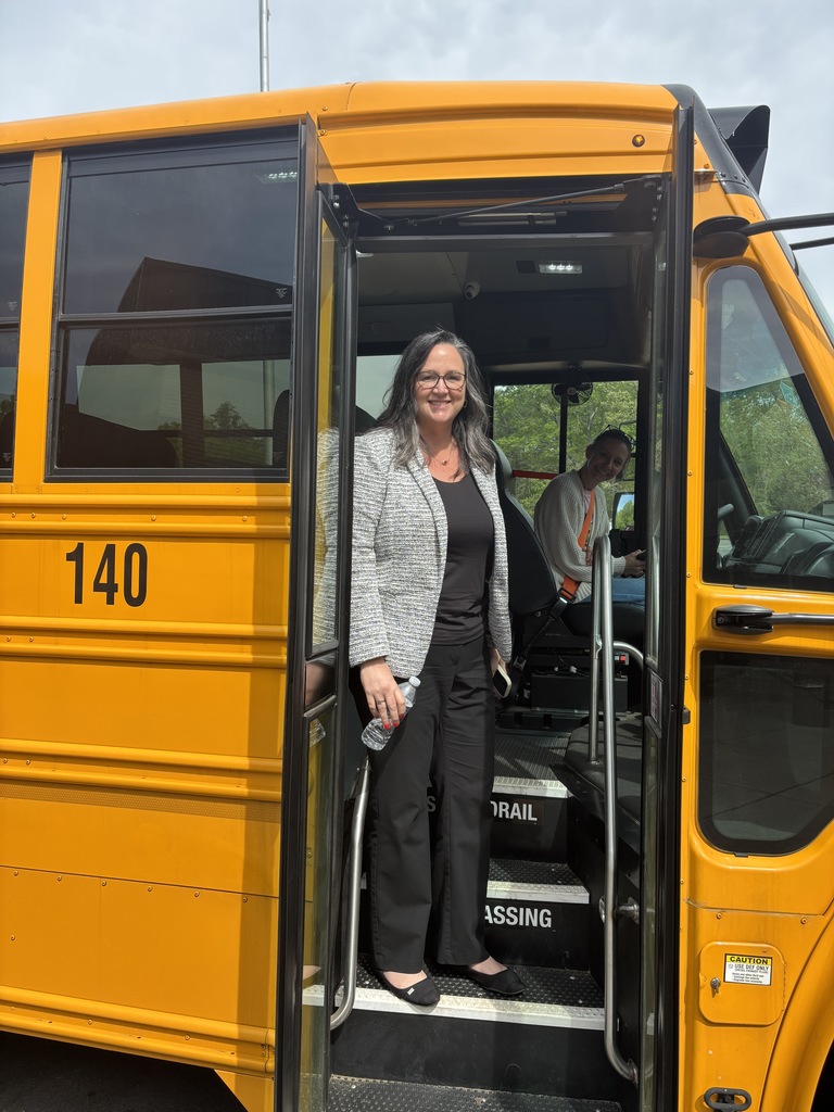 A woman wearing glasses and a gray blazer stands at the open door of a yellow bus labeled 140.