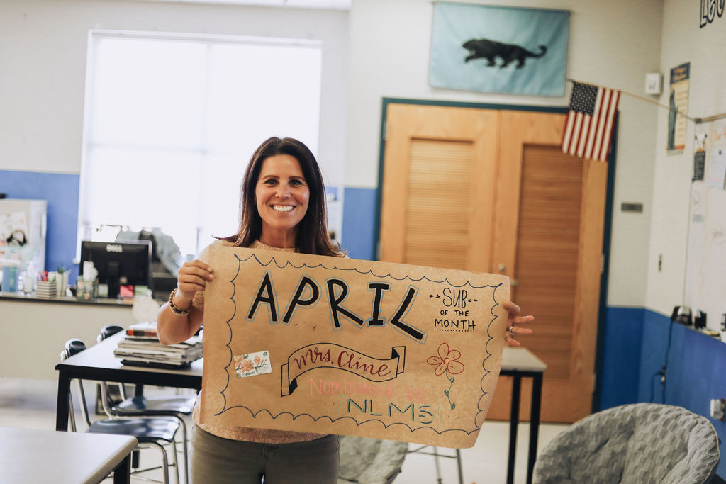 A woman in a classroom holds a cardboard sign that says "April" with stickers and decorations.