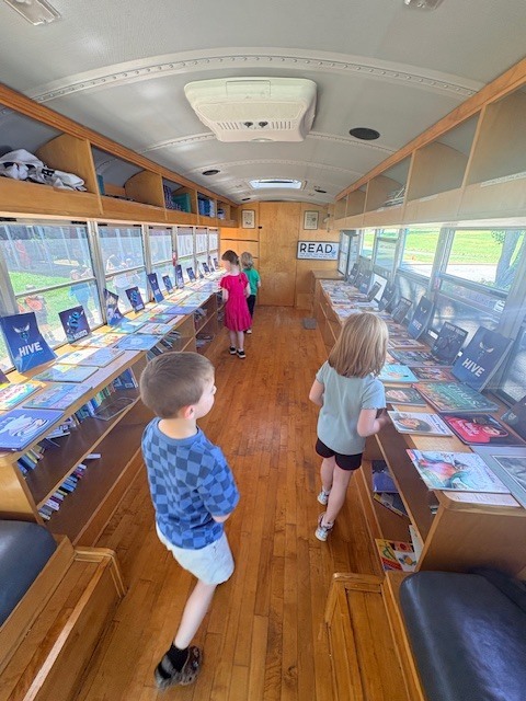 A view from inside a school bus shows kids exploring bookshelves filled with books. They are standing on a wooden floor.