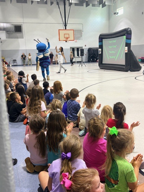 Kids are sitting on the floor, watching a mascot perform in an indoor basketball court.