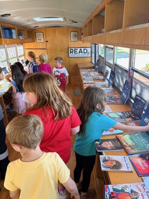Children stand in a classroom, looking at books on a table. A white sign reading "READ" is above them.