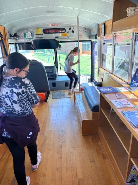 Two women inside a converted bus with wooden flooring, shelves, and an open door. One woman holds a mop.
