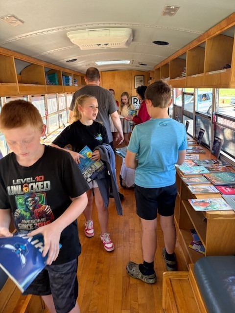 Children examine books on a wooden shelf in the interior of a repurposed bus. Shelves line the walls and ceiling.