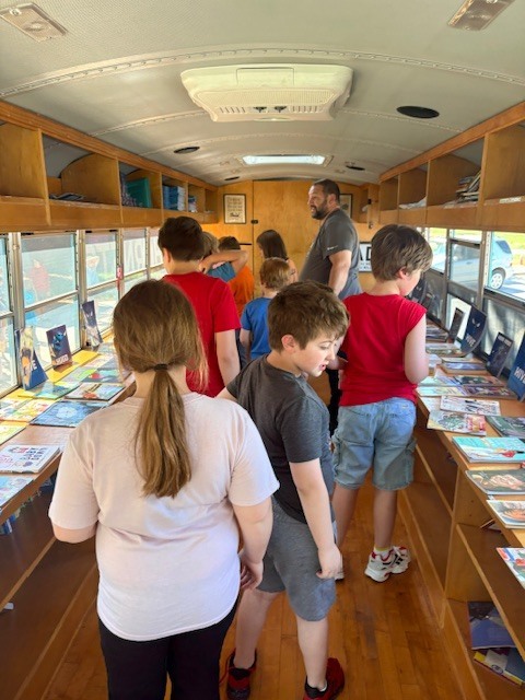 A group of children and an adult explore a book-filled bus. Books are on shelves and tables.