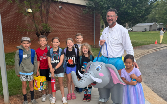 Adults and children stand on a sidewalk, a man in elephant costume, and a large tree in the background.
