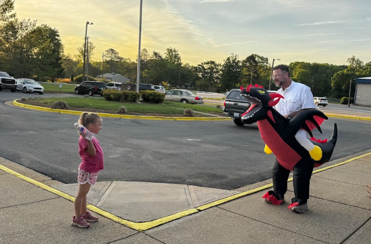 A man dressed as a dragon stands next to a young girl on a sidewalk. Trees and parked cars are in the background.