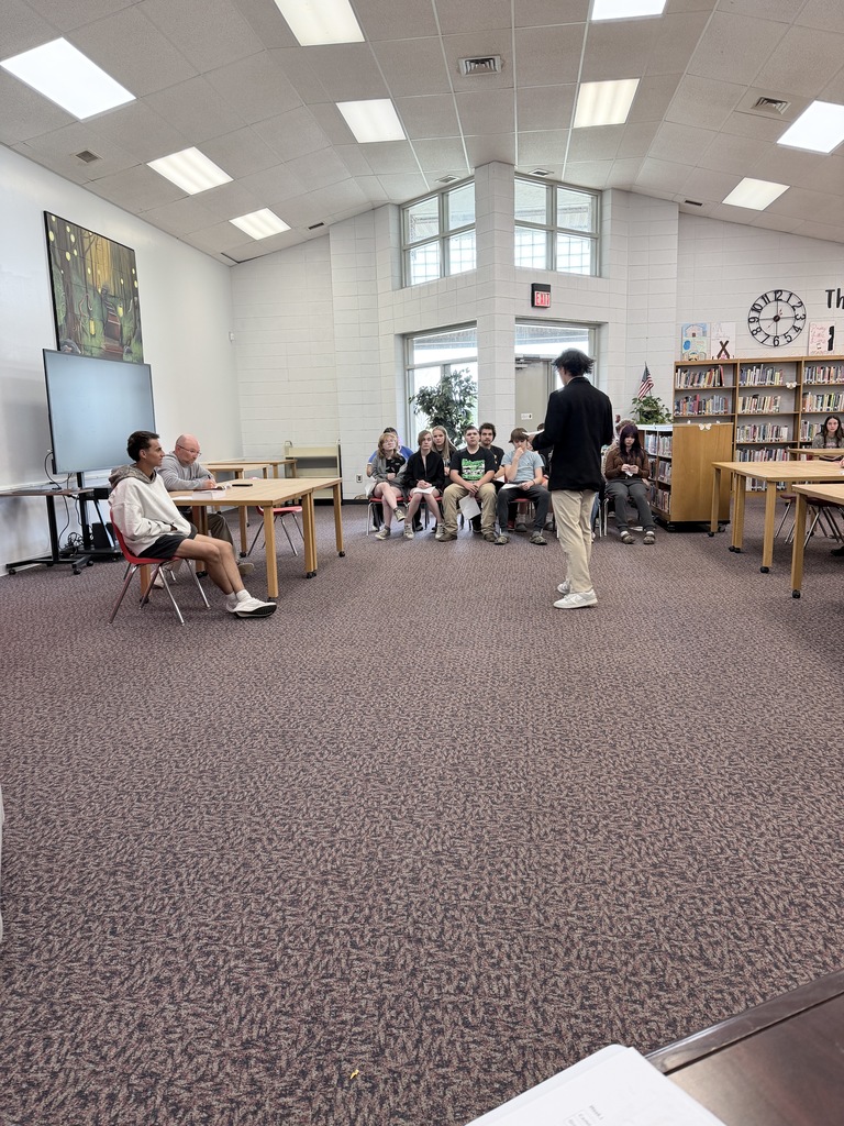 A student in a black blazer stands with their back to the camera, addressing a group of seated students in a school media center. A man and another student sit at a nearby table acting as a judicial panel.