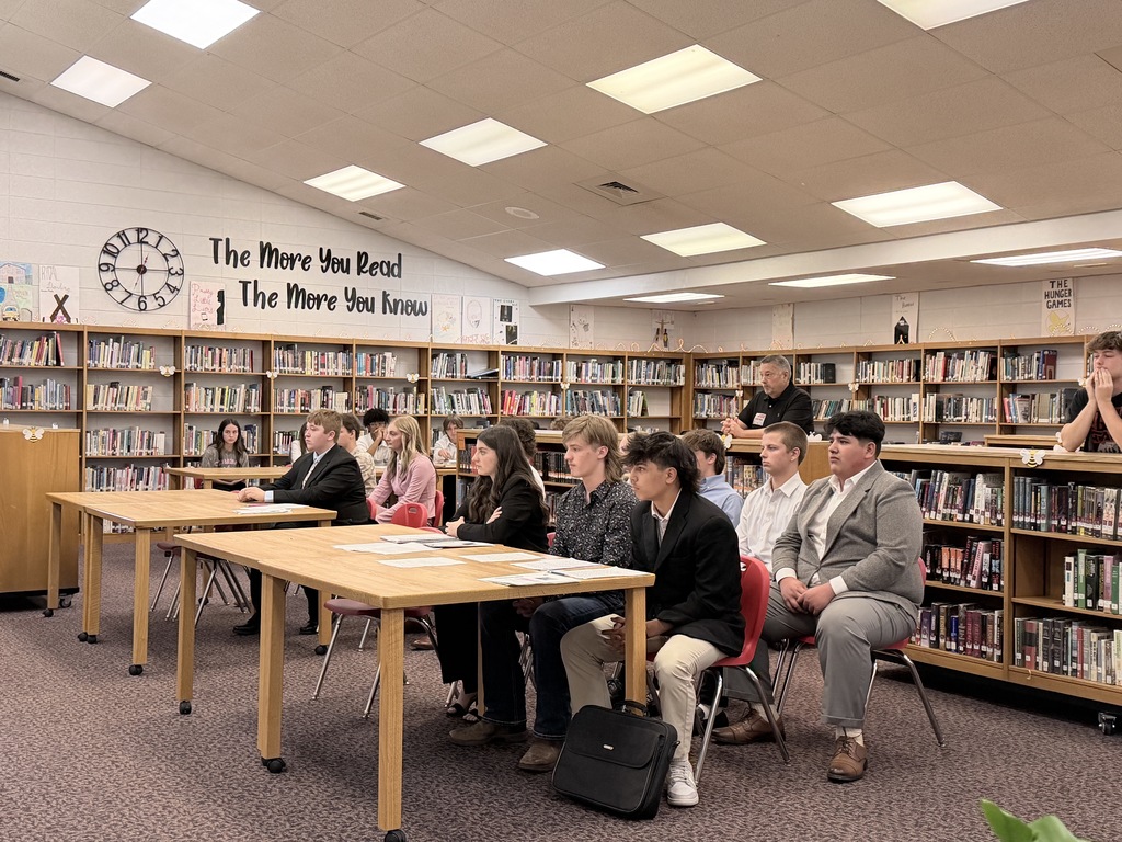 A row of students dressed in professional attire, including suits and blazers, sit at a long wooden table in a library. They are focused on the proceedings of a mock trial taking place in front of them.