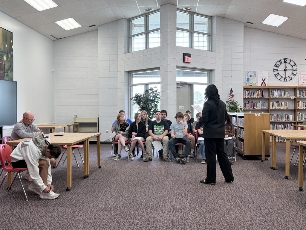 A student dressed in a professional black suit leans over a wooden table to sign or review a document. A man in a grey sweatshirt sits at the table watching, while other students observe from red chairs in the background.
