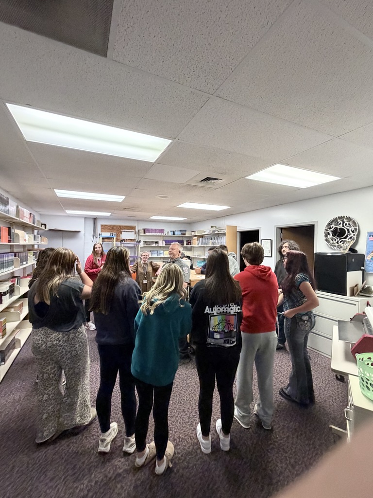 A group of people in a library with bookshelves, standing around a man speaking in front.