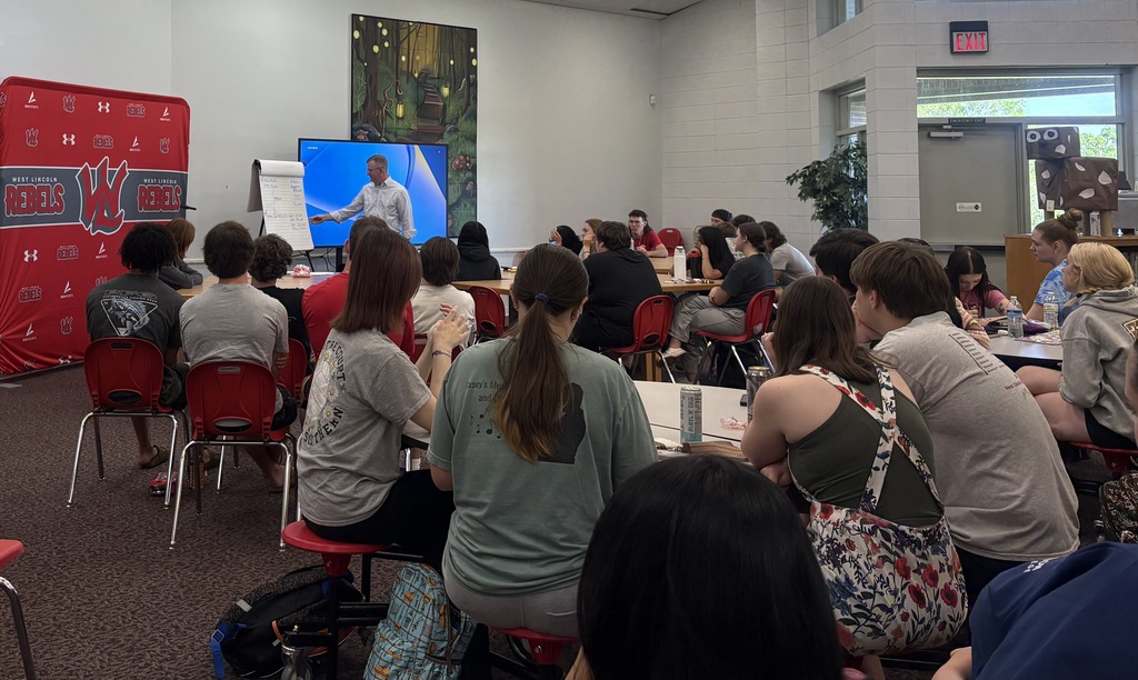 A male presenter in a light blue button-down shirt stands at the front of a room, gesturing toward a whiteboard with notes. A large digital screen behind him displays a blue desktop background. To the left is a red "West Lincoln Rebels" backdrop. A diverse group of high school students is seated at tables and in chairs, listening to the presentation. On the right, a cardboard cutout of a cow is visible near an exit door.