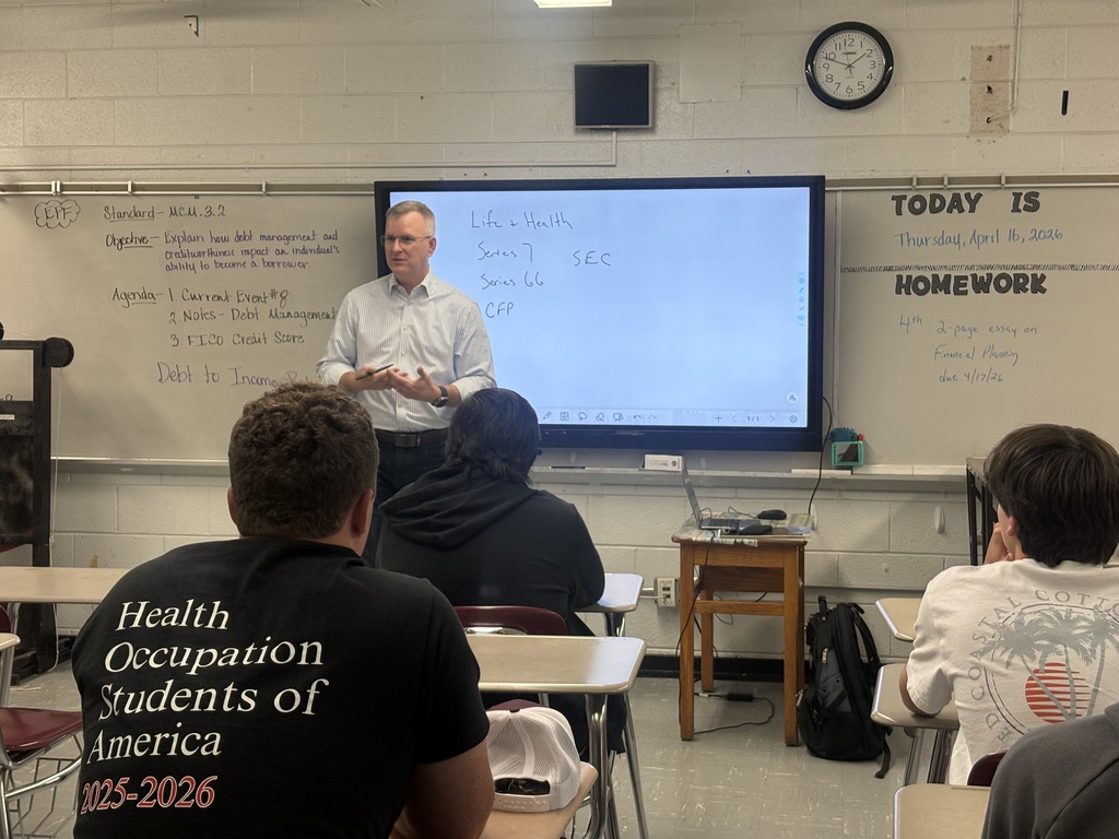 Man in a white shirt standing in front of a screen while several students look on.