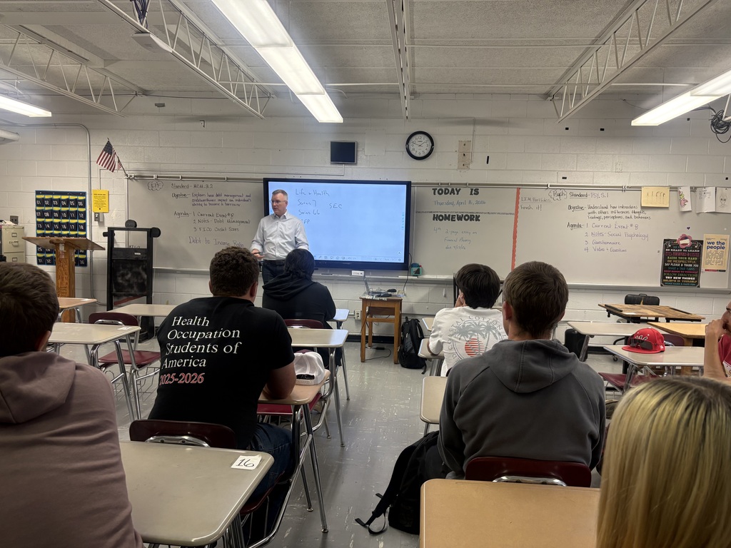 A classroom with a teacher at a podium. Students sit in rows with desks. A projector screen is mounted on a wall.