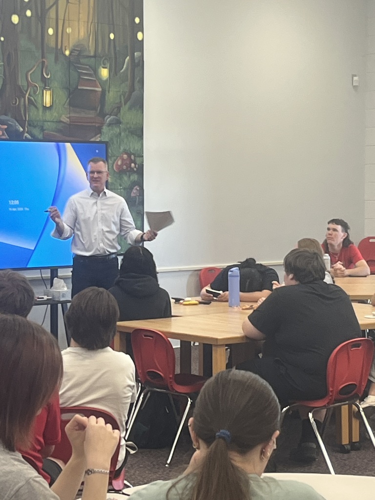 A person speaks to a group seated at tables in a classroom with a large screen behind them.