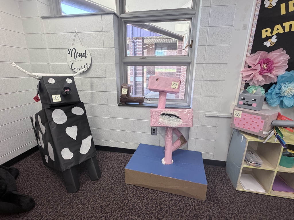 Three large cardboard sculptures are positioned in the corner of a room. Number 23 is a tall black and white spotted cow with horns. Number 24 is a tall pink flamingo standing on a blue base. Number 25 is a small brown animal on a windowsill. Number 26 is a grey and pink pixelated cat sitting on a small wooden cubby.
