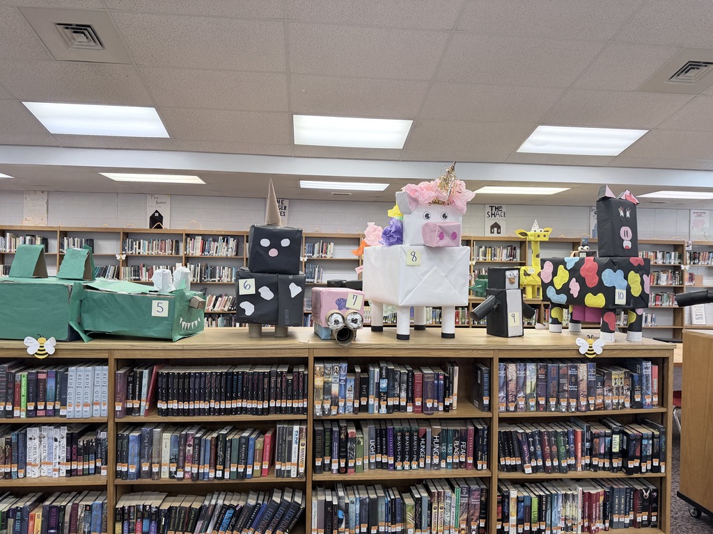 A collection of cardboard animal sculptures, numbered 5 through 10, are displayed on top of a library bookshelf filled with books. The animals include a green alligator (5), a black and white panda (6), a pink pig (7), a white unicorn with pink floral hair (8), a penguin (9), and a multi-colored spotted cow (10).