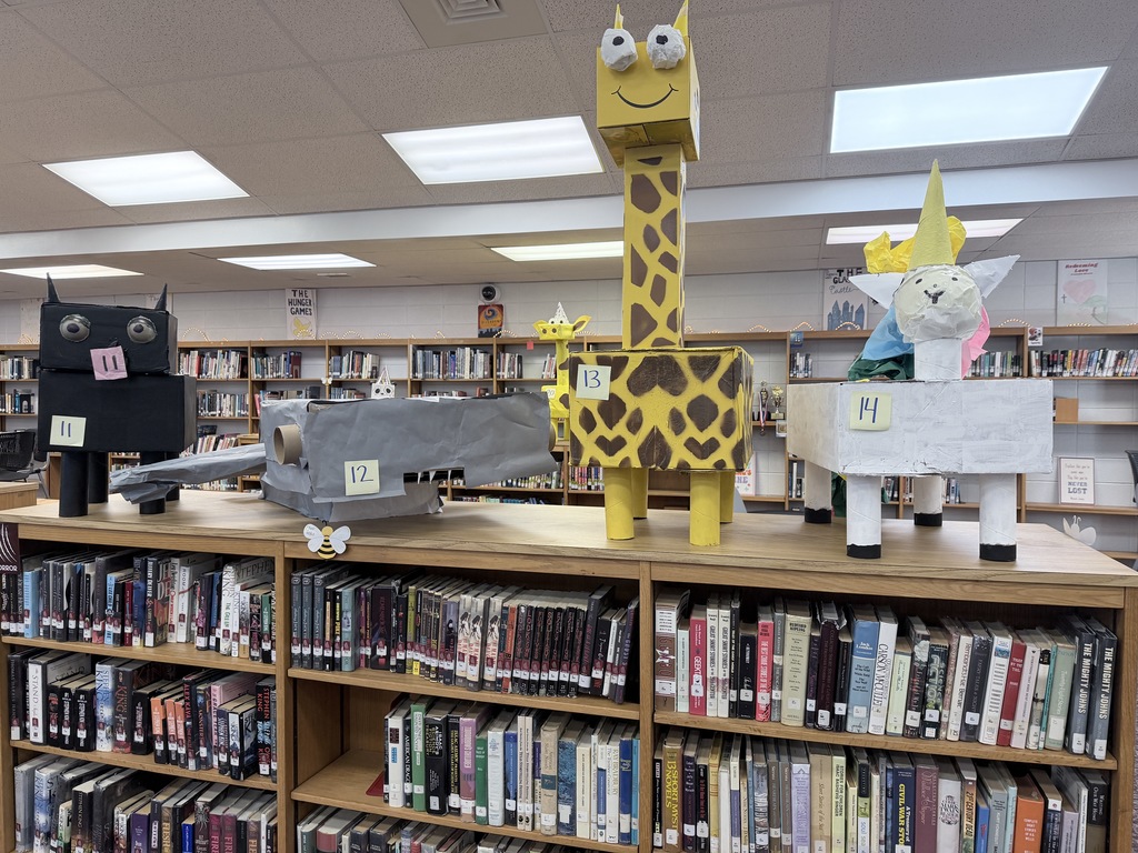 A row of student-made cardboard animal sculptures displayed on top of library bookshelves. Sculpture 11 is a black cat with large eyes; 12 is a grey shark or whale; 13 is a tall yellow giraffe with brown spots and a smiling face; and 14 is a white unicorn with a yellow horn and colorful tissue paper mane. The bookshelves below are filled with various fiction and non-fiction titles.