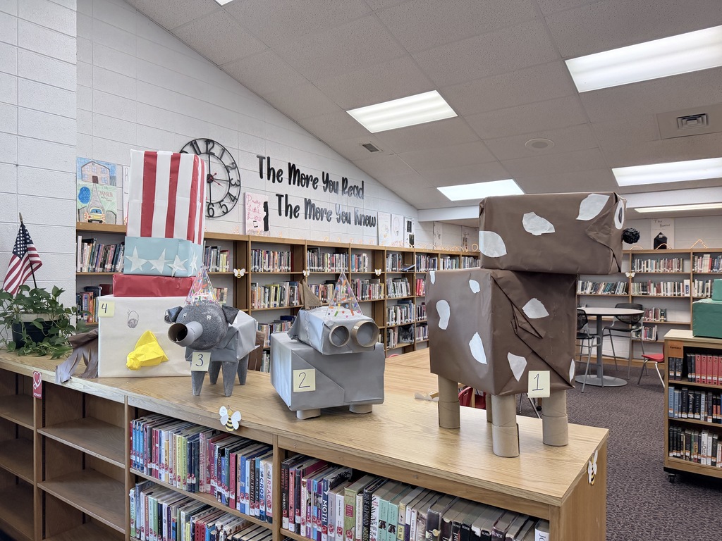 A row of student-made animal sculptures created from cardboard boxes and paper sits atop a library bookshelf. The sculptures are numbered: 1 is a large brown and white spotted cow; 2 is a grey elephant with a party hat; 3 is a grey rhinoceros with a party hat; and 4 is a white chicken with a red and white striped hat. A sign in the background reads, "The More You Read, The More You Know."