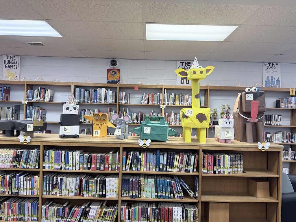 A library bookshelf features student-made cardboard animal sculptures numbered 15 through 22. The display includes a black animal (15), a panda (16), an orange cat (17), a grey mouse (18), a green creature (19), a tall yellow giraffe with brown spots (20), a small white cat (21), and a large brown dog with a long pink tongue (22).