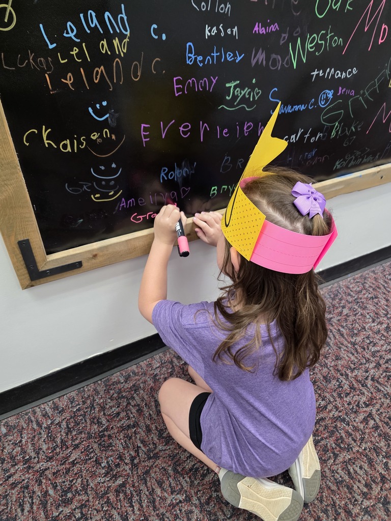 A child in a yellow cone hat writes on a chalkboard filled with various names and drawings.