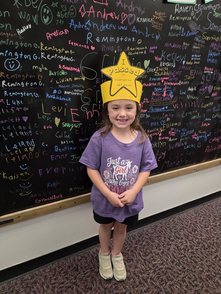 A young child with a yellow star crown stands in front of a blackboard filled with writing.