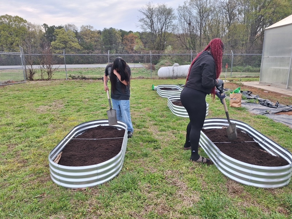 Two people are working in raised garden beds with shovels, one with curly hair, another with a ponytail.